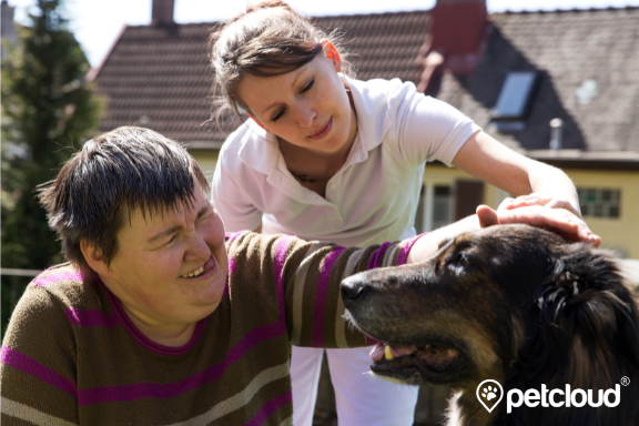 A woman in a striped sweater smiles while petting a large dog, assisted by another woman in a white shirt. They are outdoors in a garden, with houses and a tree in the background. The scene exudes warmth and companionship, highlighting animal assisted therapy in Australia. The "petcloud" logo is in the bottom right corner.