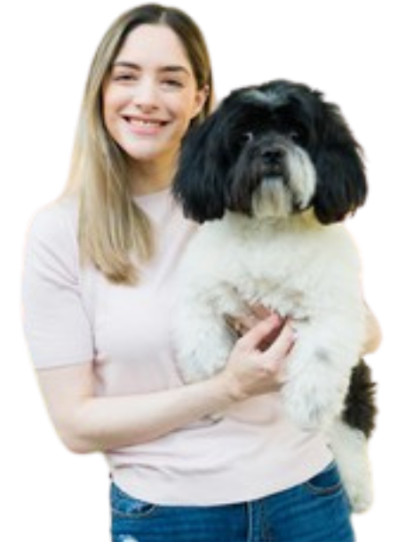 A person with long, light brown hair, dressed in a light pink top and blue jeans, is smiling and holding a small black and white dog. The background is not present in the image.