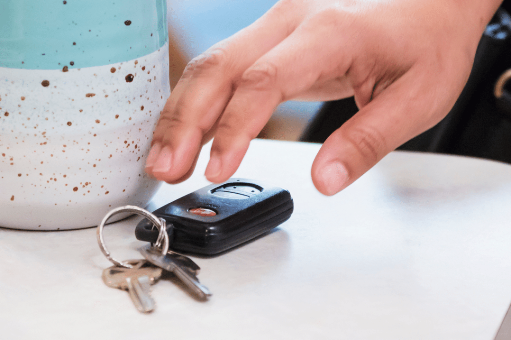 A close-up image of a hand reaching for a set of keys and a car key fob placed on a light-colored surface near a speckled ceramic cup with a blue and white gradient.
