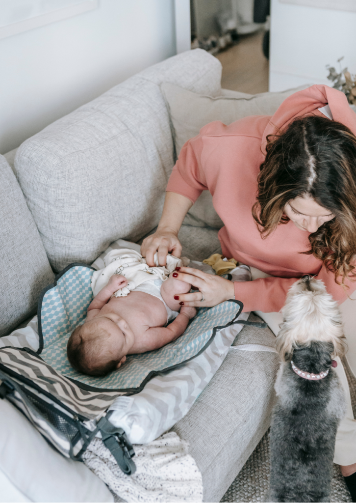 A woman dressed in a pink top is gently attending to a baby lying on a changing mat on a gray couch. A small dog is standing near her, looking up attentively. Various baby items are scattered around.