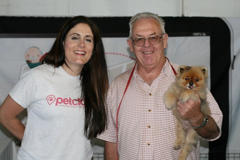 A woman and an older man stand smiling. The woman, wearing a white 'petclix' t-shirt, has long, dark hair. The man, wearing glasses and a checkered shirt, holds a small, fluffy brown dog. They are indoors, with a banner partially visible in the background.