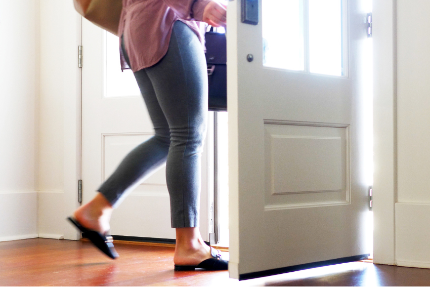 A person with a brown bag steps through a partially open door into a brightly lit room. The individual is wearing gray leggings, black slip-on shoes, and a light-colored top. Sunlight streams through the door, casting a warm glow on the scene as they check their phone for a pet sitter booking confirmation.