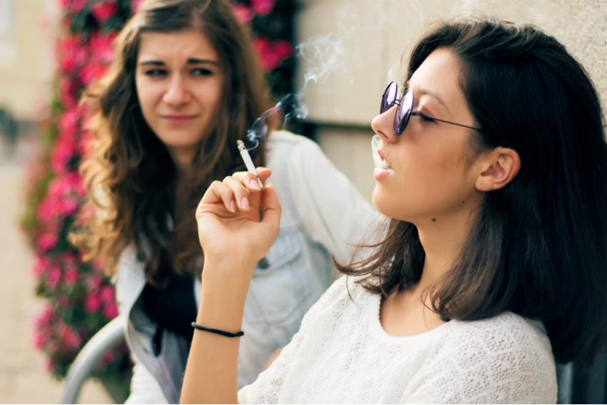 A young woman smokes a cigarette while wearing sunglasses and leaning against a wall. Another young woman in the background, with wavy hair and a denim jacket, looks at the smoker with a disapproving expression. Pink flowers surround them as if witnessing a forgotten pet sitter booking.