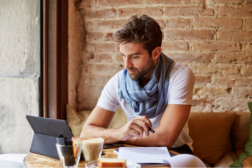 Man sitting at a cafe table, wearing a scarf and holding a pen, looking at a tablet with the logo of his dog walking business. There are documents, a cup of coffee, and a slice of cake on the table. The background features an exposed brick wall.