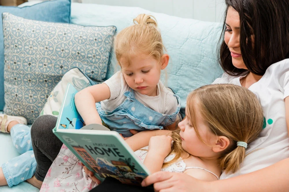 A woman is sitting on a couch with two young girls. One girl is holding a book titled "A Place Called Home" and reading it aloud. They all appear engaged and comfortable, surrounded by soft blue and white pillows, creating an atmosphere that feels like the gentle embrace of home care services.