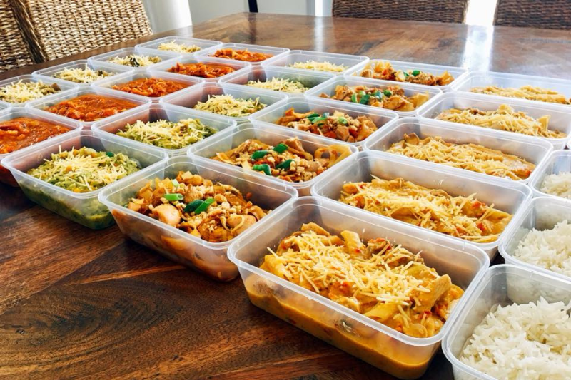 A large wooden table covered with rows of plastic containers filled with a variety of prepared meals, including pasta dishes topped with grated cheese, rice, green beans, and other colorful ingredients, all organized neatly for meal prepping as part of comprehensive home care services.