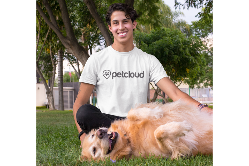 A smiling person wearing a "petcloud" t-shirt sits on grass with a happy assistance dog, a golden retriever, lying on its back. Trees and a building are visible in the background.