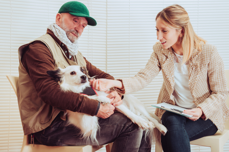 A man in a green cap and light brown jacket holds a white and black dog while a woman in a beige plaid jacket sitting next to him pets the dog and smiles. The woman, engaged in the recovery of PTSD, has a clipboard and pen in her other hand. They are in a room with white blinds.