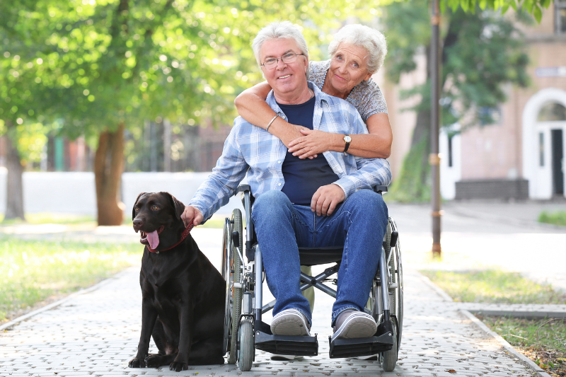 A smiling man with PTSD in a wheelchair with a dog by his side, holding its collar, and being cuddled by a woman from behind.  
