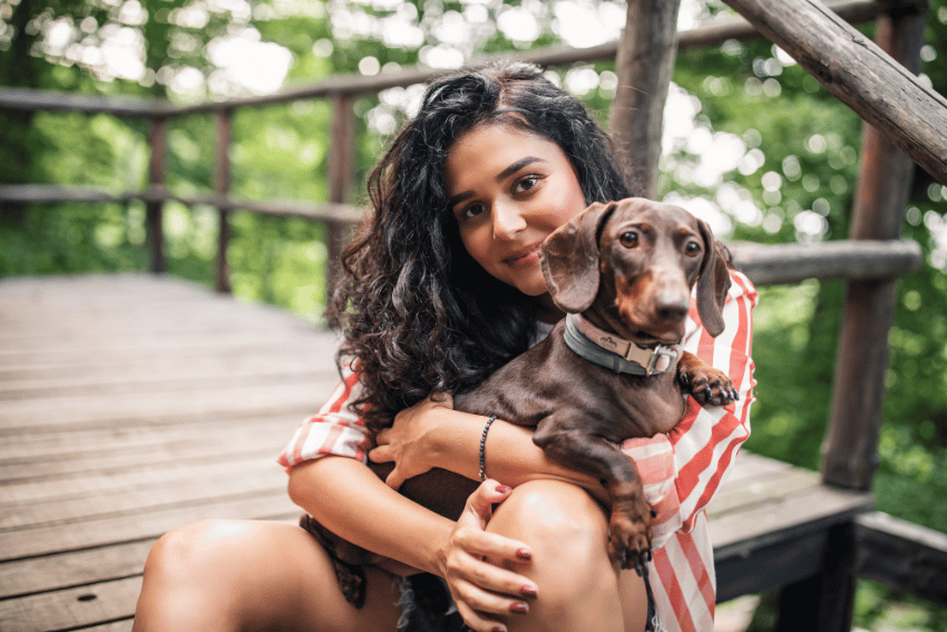 A female Melbourne University student with long, curly dark hair, wearing a red and white striped shirt, sits on wooden steps outdoors in a lush, green forest setting. Hugging a small brown dachshund and smiling at the camera, she seems tranquil amidst nature. Her serene expression amidst this peaceful scene is reminiscent of the focus Melbourne Uni students often bring to their search for job opportunities.