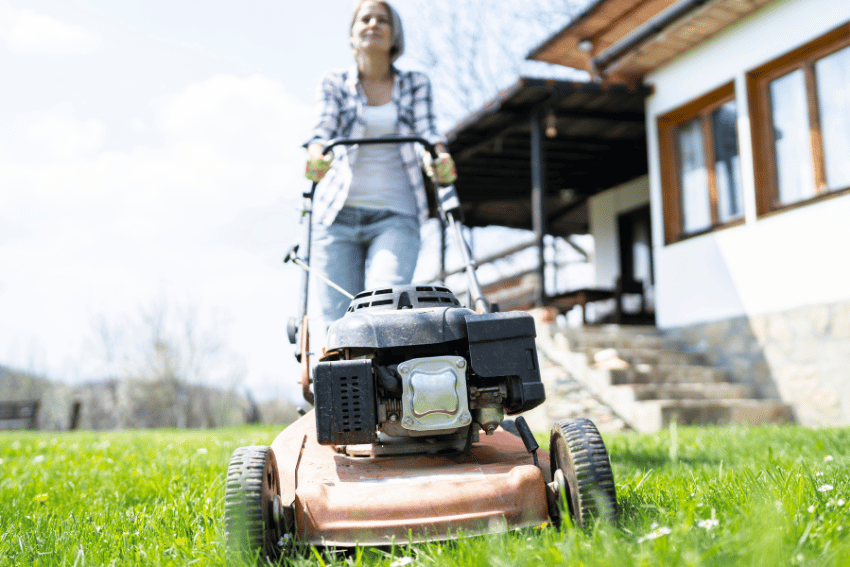 A person is safely guiding a lawnmower across the grassy lawn in front of a house on a sunny day. The porch with several large windows overlooks pets lounging in the shade, while the person goes about their task in casual clothing.