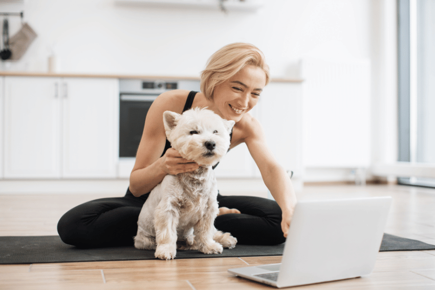 A woman with short hair sits on a yoga mat in a modern kitchen, smiling at a laptop. A small white dog sits beside her, also facing the laptop. The bright and minimalistic space echoes the easy living of the Sunshine Coast lifestyle and Affordable Long-Term Pet Boarding on the Sunshine Coast