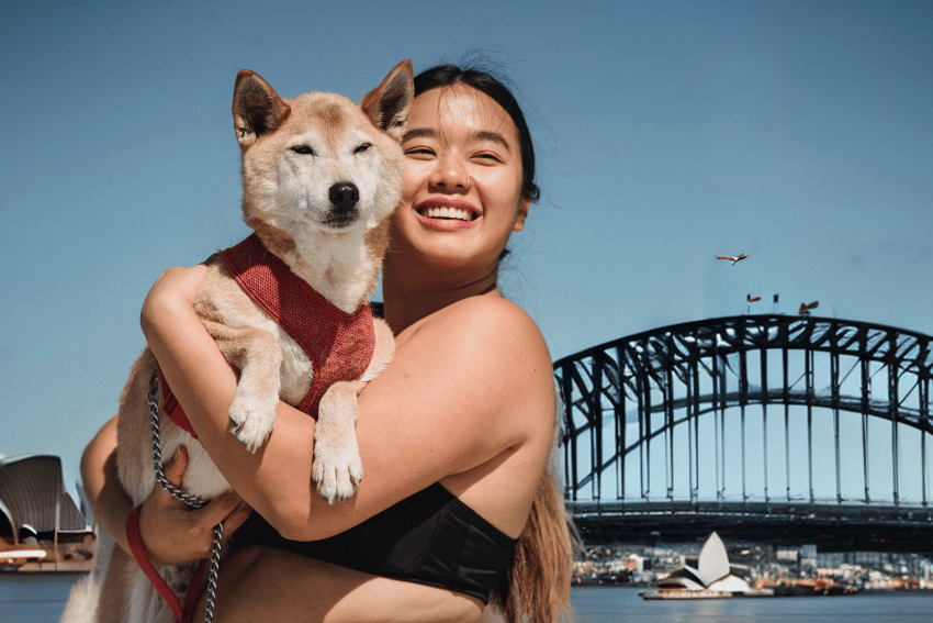 A woman beams with joy, holding her dog in front of the iconic Sydney Opera House and Harbour Bridge. The dog sports a red harness, adding charm to the picturesque scene. As small planes glide through the clear blue sky, one can't help but think this city offers the best dog boarding in Sydney.
