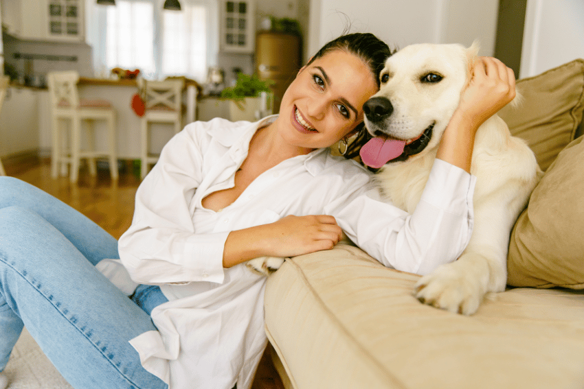 A woman in a white shirt and jeans sits on the floor, leaning against a beige sofa, smiling beside her trusted dog. The background reveals a bright, modern kitchen from their cozy home in Melbourne.