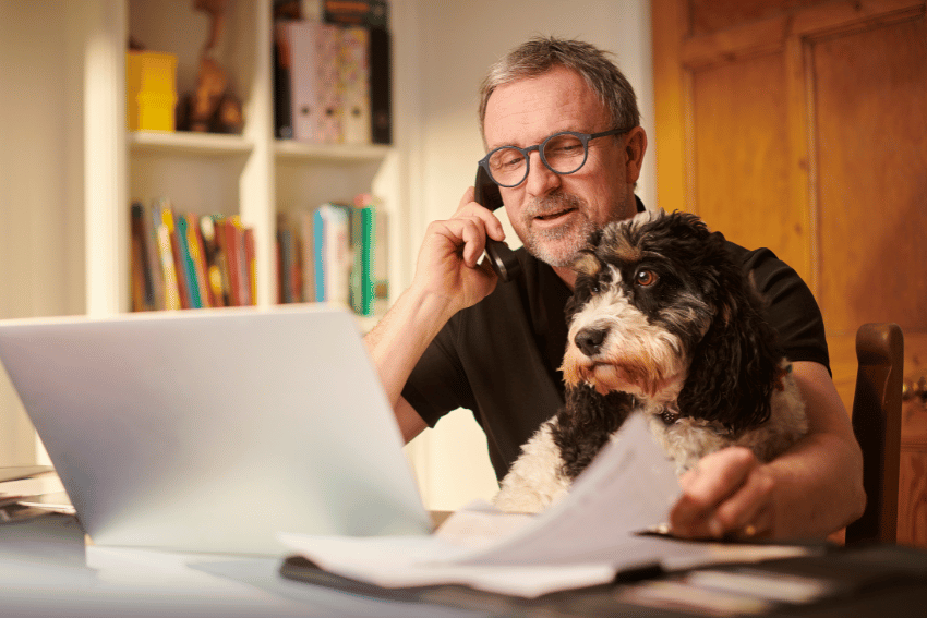A man with glasses sits at a desk, talking on the phone and looking at a laptop while holding papers. His black and white dog rests on his lap in a home office, perhaps seeking advice from cheap vets that recommend home remedies.