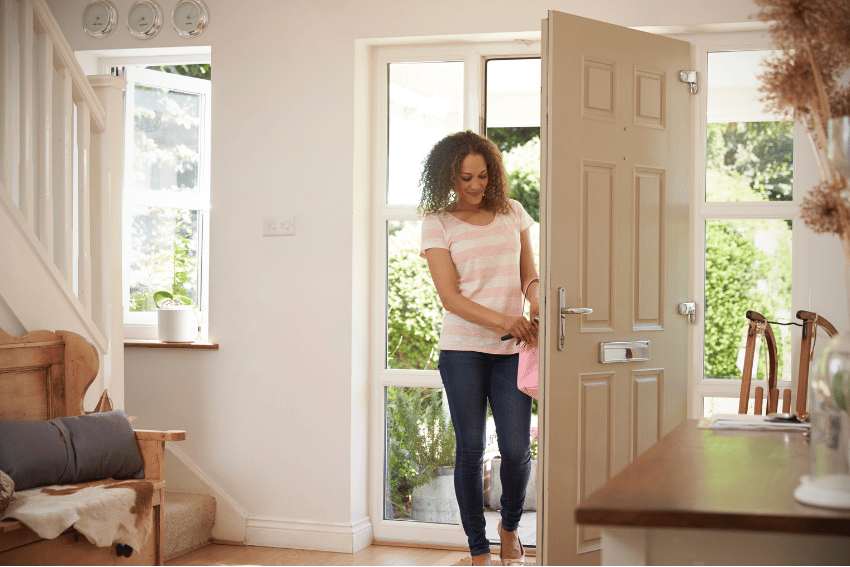 A woman stands at an open front door in a bright, tidy hallway, holding a pink bag and looking down as sunlight streams in through the windows behind her, perhaps thinking about Sick Leave for Pets Australia.