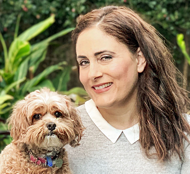 Deb Webber with long brown hair and a white-collared gray sweater smiles while holding a small, curly-haired brown dog in a garden with green plants in the background.