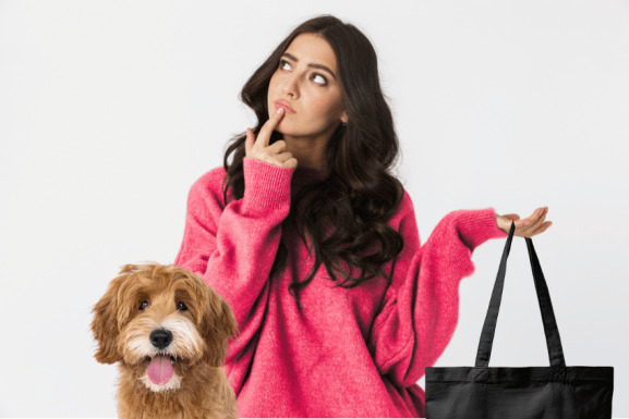 A woman in a bright pink sweater looks up thoughtfully with a finger on her lips, holding a black tote bag—perhaps pondering how to prepare your dog for a pet sitter. A fluffy brown dog with a happy expression sits in front of her.