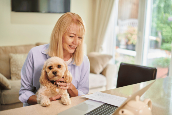 A smiling woman with blonde hair sits at a table with a small dog on her lap, researching PetCloud vs Pawshake—which is the safer choice for Australian pet owners—on her laptop. Papers and a piggy bank rest nearby in her cozy living room.