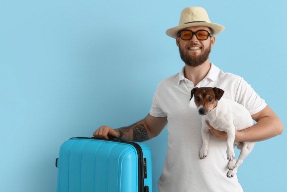 A smiling man in a white shirt, sunglasses, and a hat stands next to a blue suitcase, holding a small dog in his arms against a light blue background- ready for travel after checking the latest pet sitter cost Australia guide.